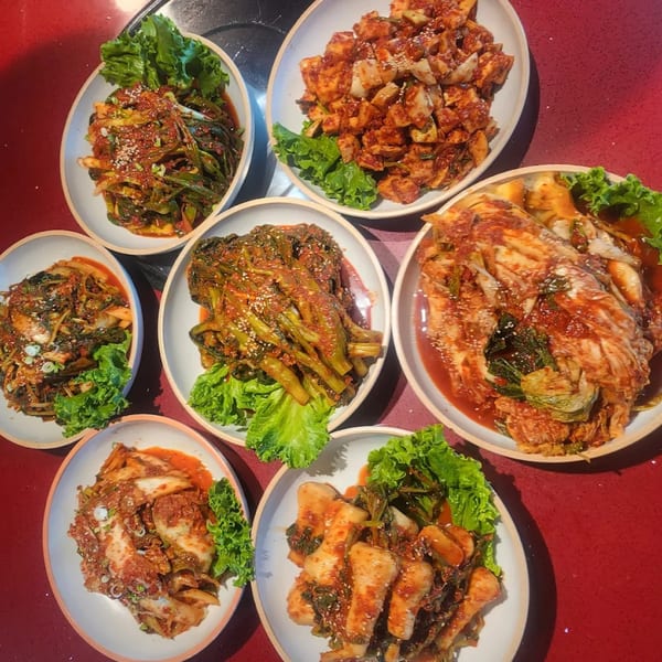 An overhead view of seven white bowls filled with various types of vibrant red Korean kimchi on a red table.
