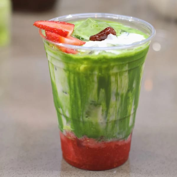A vibrant green and red layered drink with strawberries and cream in a clear plastic cup.